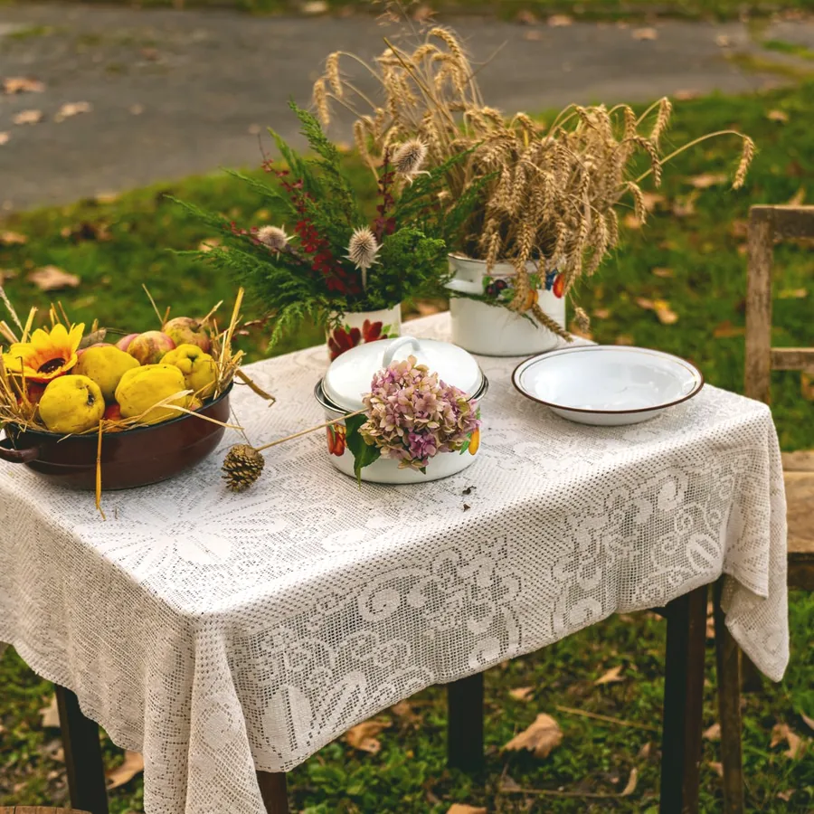 A harvest table on open grass: a wooden bowl of quince, cut wheat, and enamel cookware on a lace cloth.