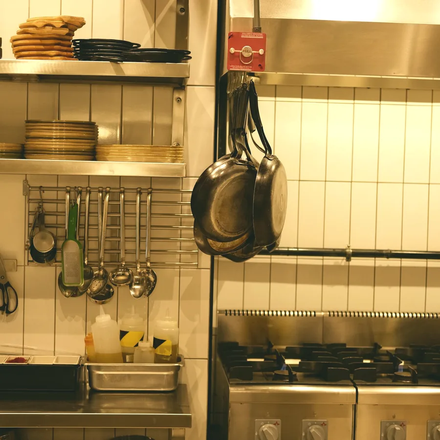 A working commercial kitchen: pans hanging above a stainless range, stacked plates on the shelf above.