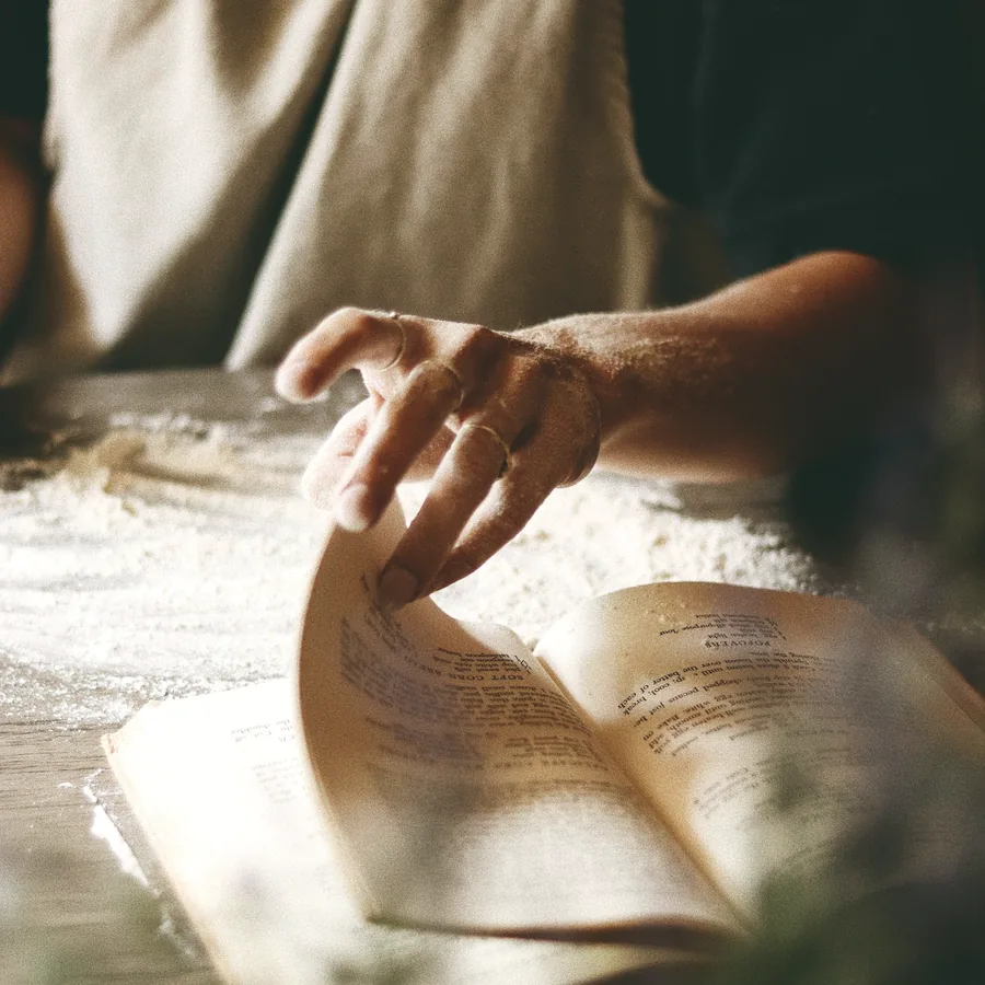 Flour-dusted hands turning the pages of an open recipe book on a wooden table.